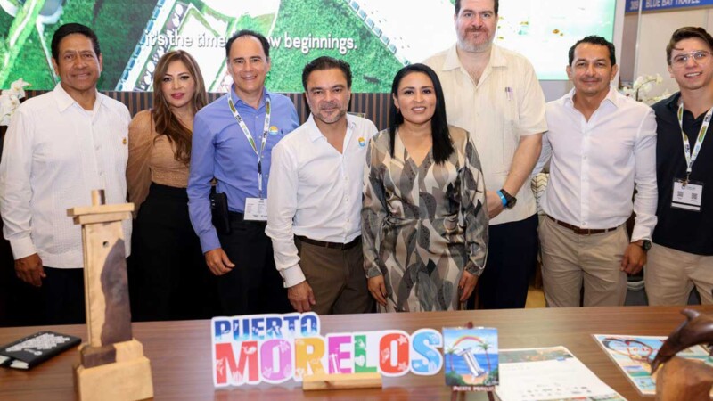 A group of professionals poses for a photo at an event, showcasing a colorful sign that reads "Puerto Morelos."