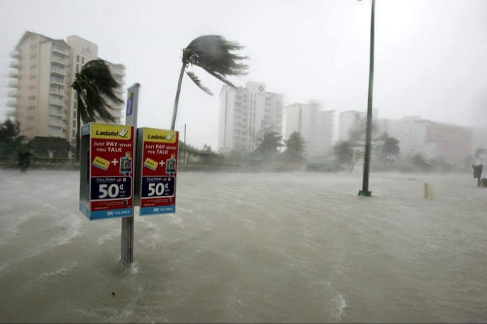 A telephone booth partially submerged in floodwaters with strong winds and palm trees bending, while buildings loom in the background under stormy conditions.