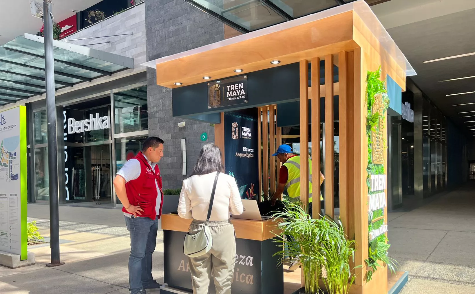 A small wooden information booth for the Tren Maya project, with a person interacting with a staff member. The exterior features greenery and signage about archaeological wealth. Nearby is a shopping area with the name "Bershka" visible.$# CAPTION