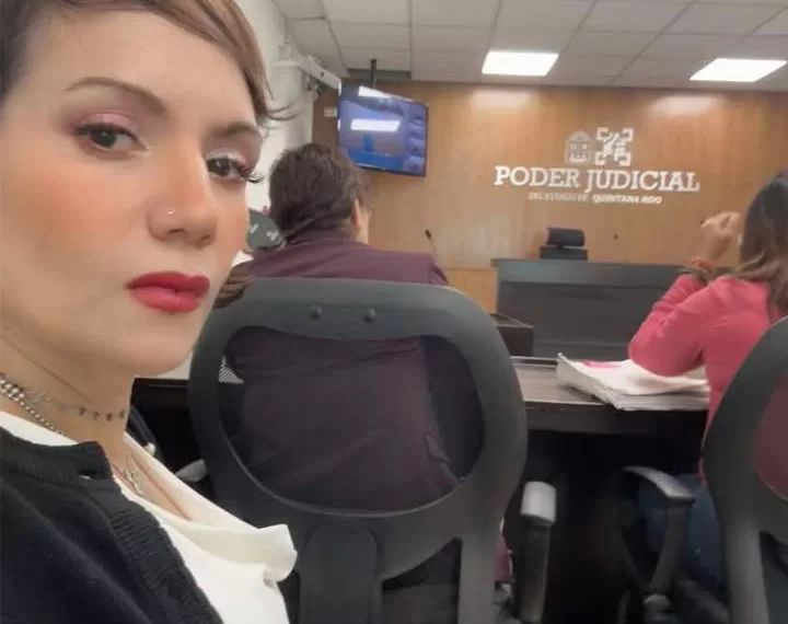 A woman with short hair and red lipstick sits in a courtroom, looking towards the camera while others are visible in the background.