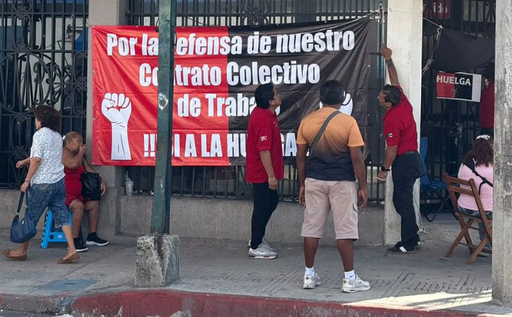 A group of people is hanging a large banner that advocates for the defense of collective labor contracts. The setting appears to be a street with several onlookers.
