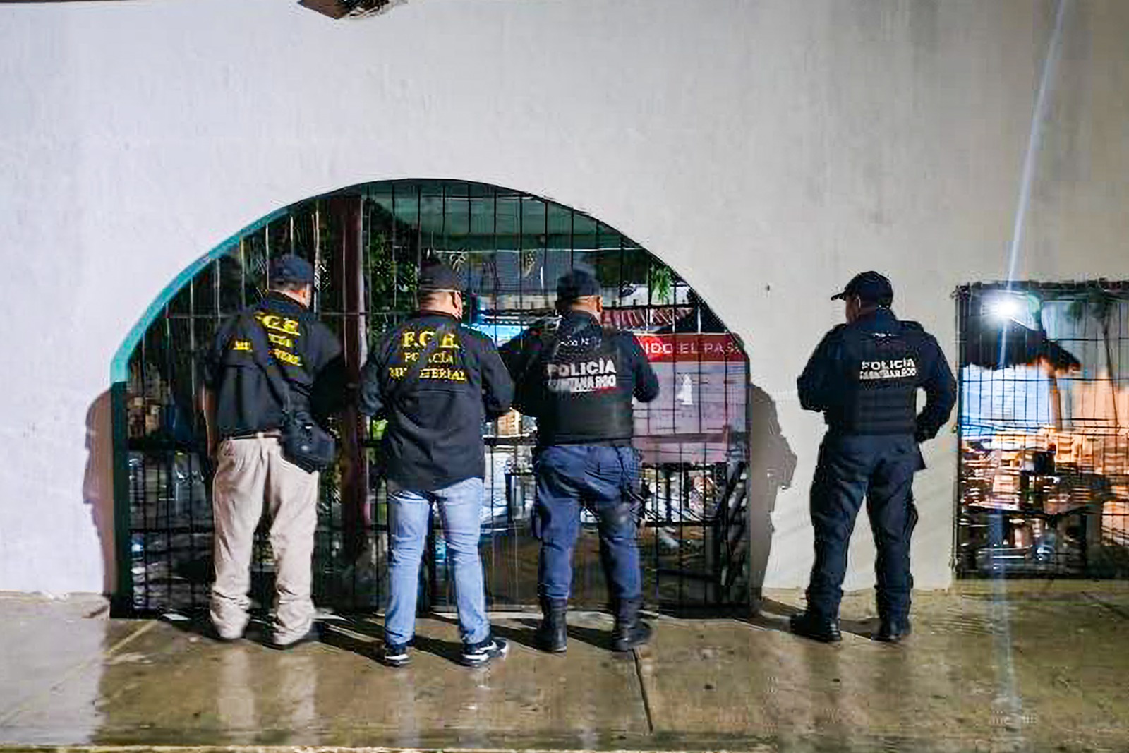 Four law enforcement officers are standing in front of a gated entrance, observing the area at night.