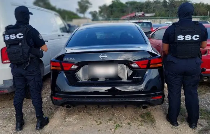 Two law enforcement officers in tactical gear standing behind a black Nissan Altima parked among other vehicles.$# CAPTION