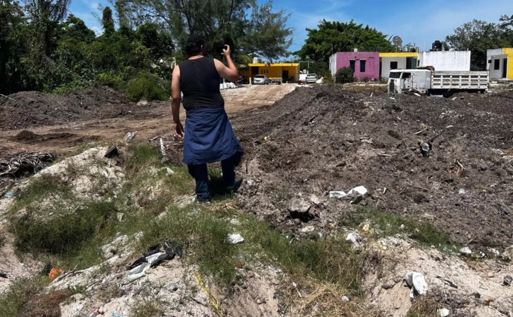 A man wearing a black tank top and blue shorts walks away from the camera in a debris-filled landscape with small hills of dirt and vegetation. A few buildings in the background can be seen, including a yellow and a pink structure. The sky is partly cloudy.