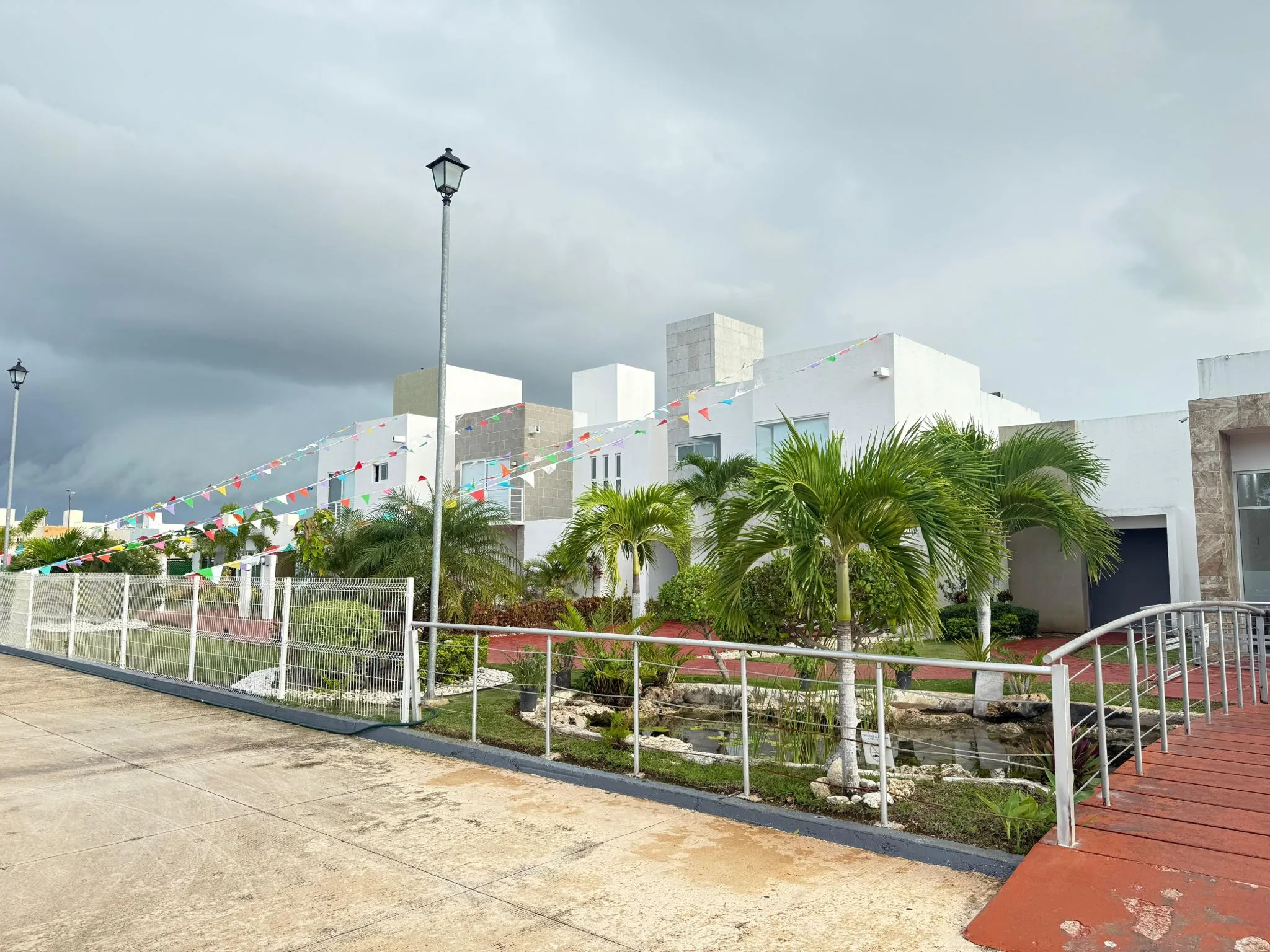 A modern residential complex featuring white buildings, lush greenery, and colorful decorative bunting under a cloudy sky.$#$ CAPTION
