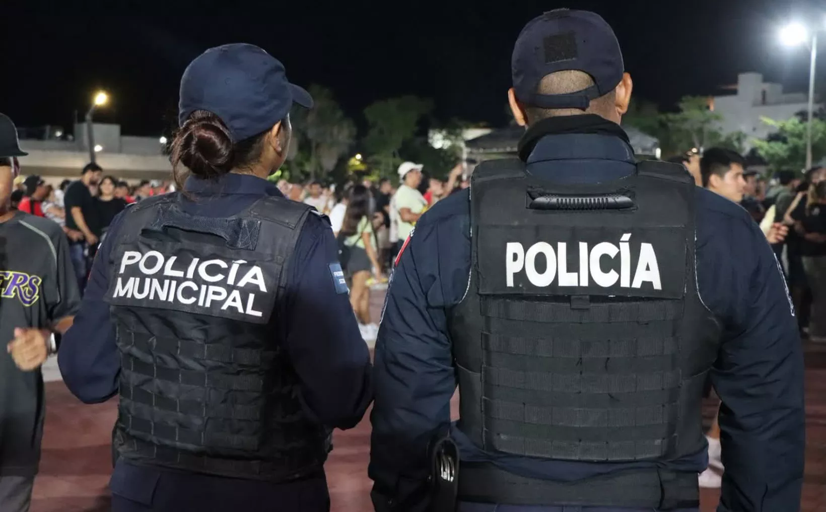 Two municipal police officers stand with their backs to the camera, focusing on an event with a crowd in the background at night.$# CAPTION