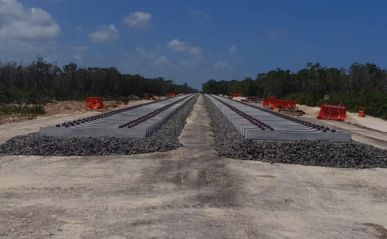 An expansive view of railway tracks under construction, with concrete sleepers and gravel on either side, set against a blue sky and green trees in the background.$# CAPTION