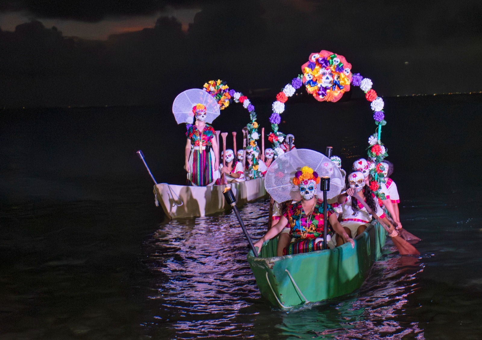 Colorfully dressed participants in traditional outfits and decorative skull masks paddle boats adorned with flowers at night.$# CAPTION