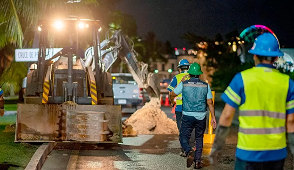 A team of construction workers is engaged in nighttime road work, utilizing heavy machinery to manage an excavation site while ensuring safety with proper gear and lighting.