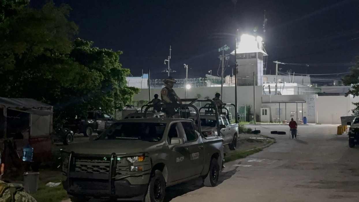 Military trucks and personnel patrolling outside a correctional facility at night. The facility is illuminated by floodlights in the background.
