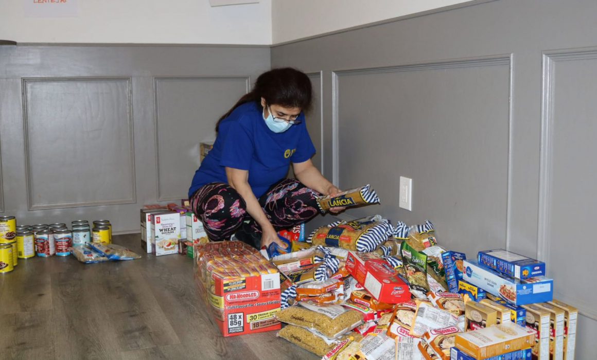 A woman wearing a blue shirt and a mask is sorting through various food items on the floor, including canned goods and packaged foods, in a community space.$# CAPTION