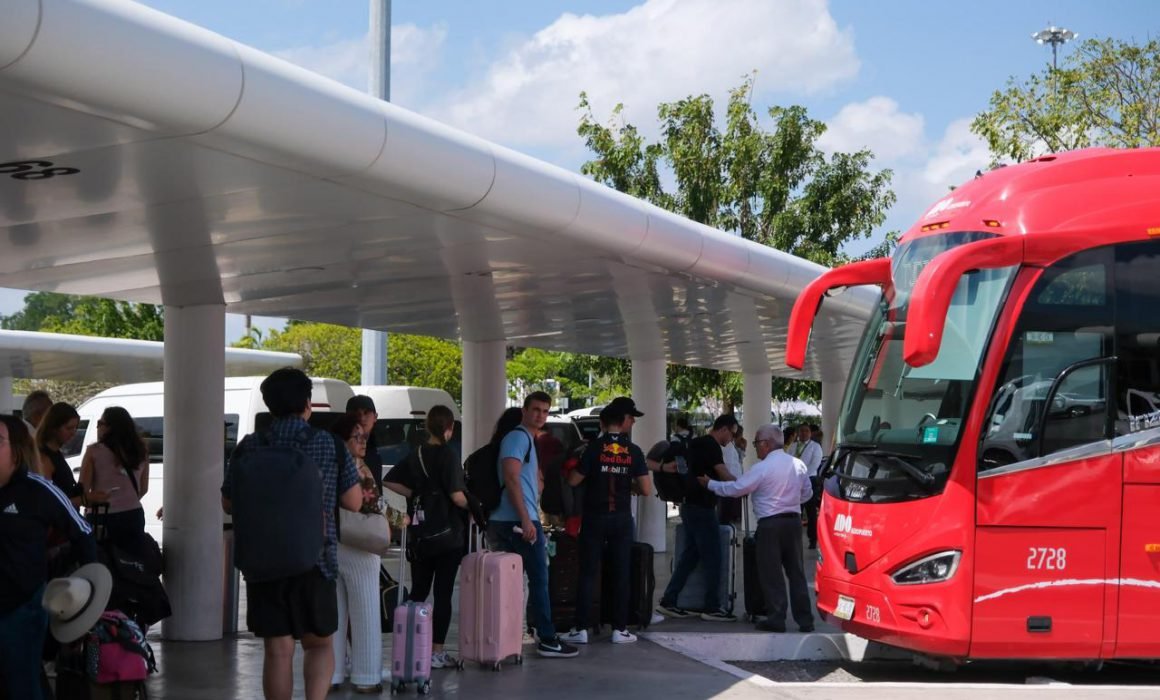 A group of passengers with luggage lined up at a bus terminal under a shaded area, with a red bus in view.$# CAPTION