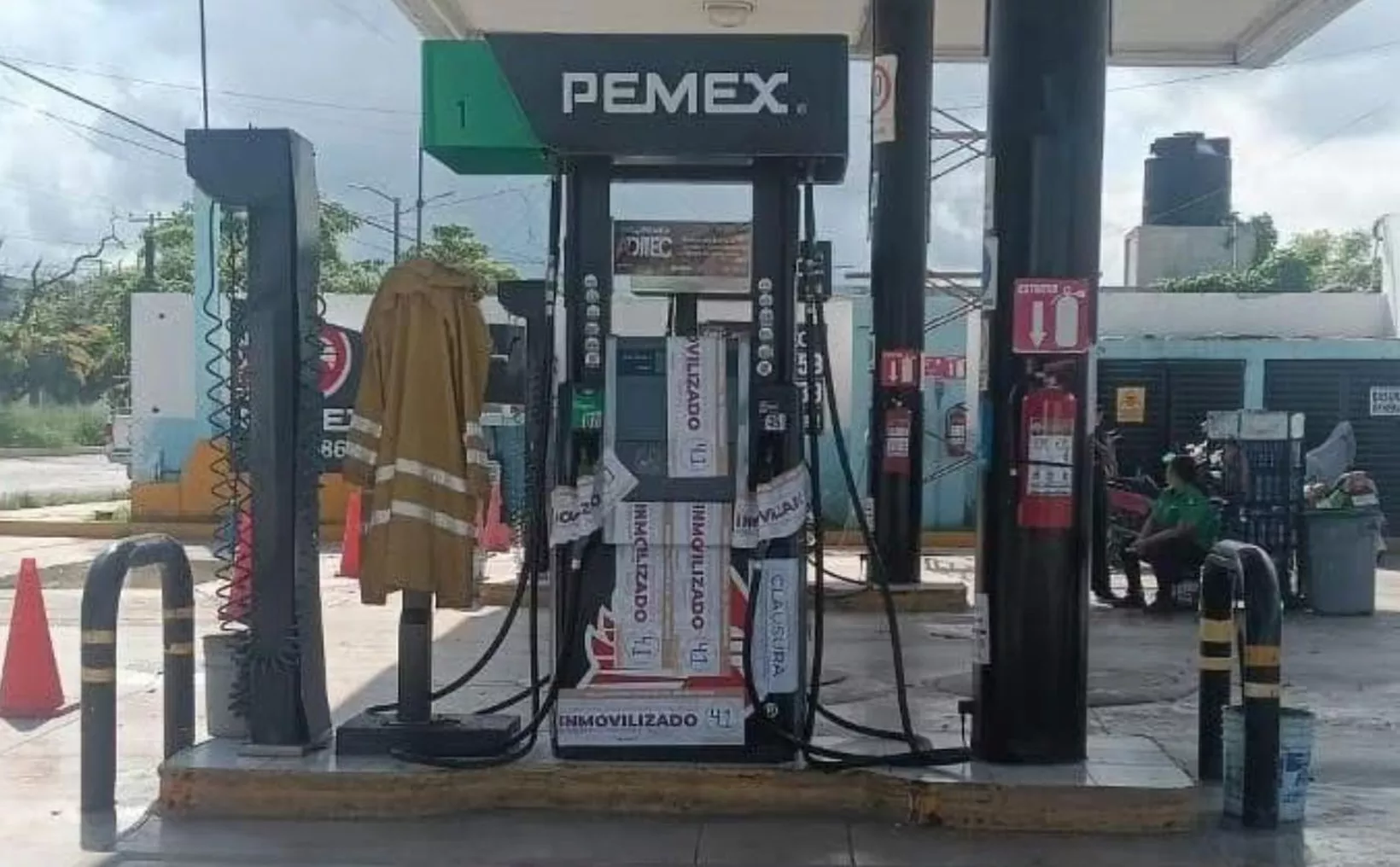 A Pemex gas station with fuel pumps, a fireman's coat hanging, and a barrier in the foreground. A person is seated nearby.