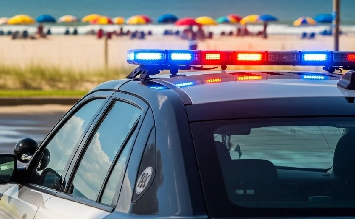 A police car with flashing lights parked near a beach with colorful umbrellas in the background.$# CAPTION
