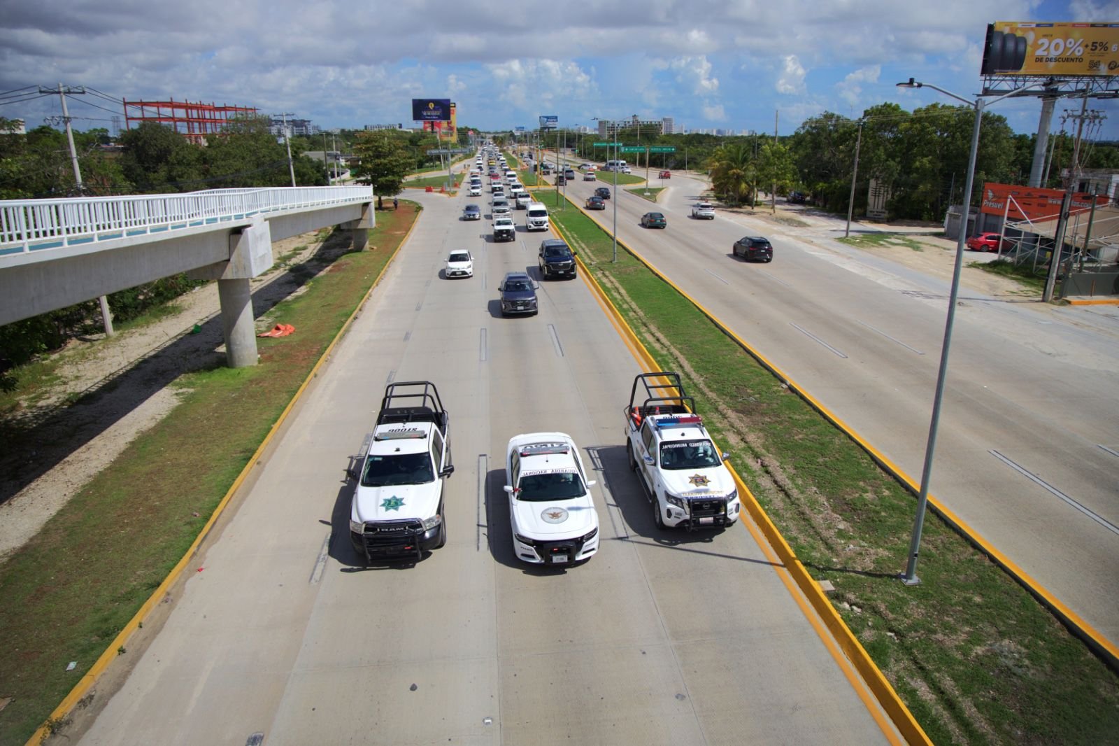 Three police vehicles and several cars traveling on a busy road under a clear sky with clouds.
