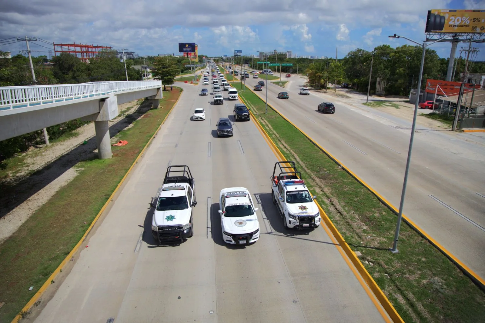 Three police vehicles and several cars traveling on a busy road under a clear sky with clouds.