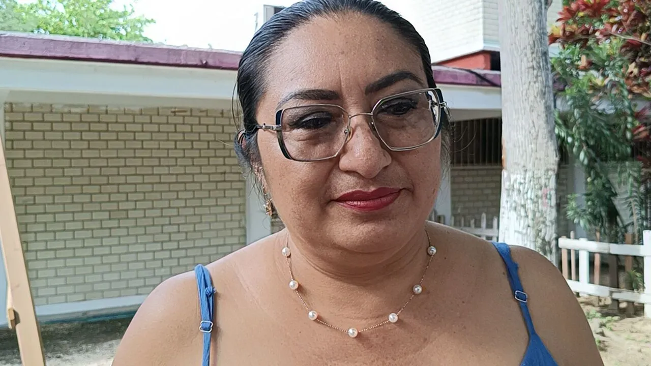 A woman wearing glasses and a necklace, with a serene expression, standing outdoors with a blurred background of trees and a building structure.$