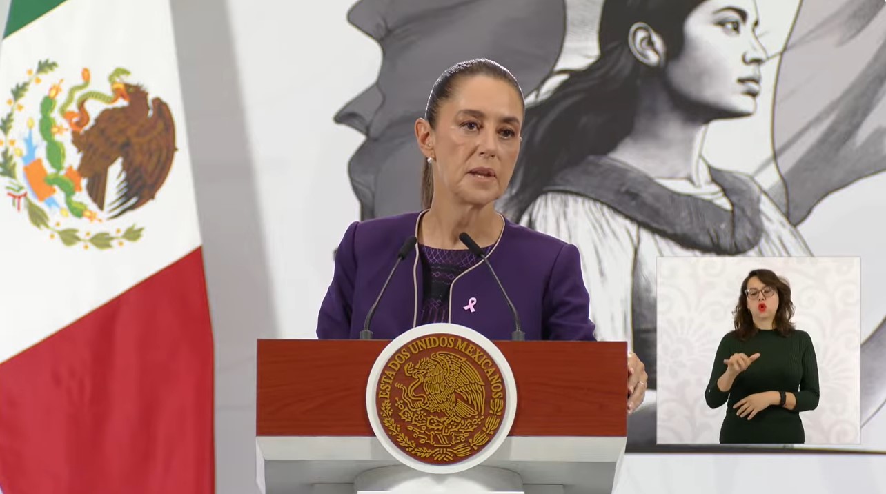 A woman in a purple suit speaks at a podium during a press conference, with the Mexican flag and a mural in the background