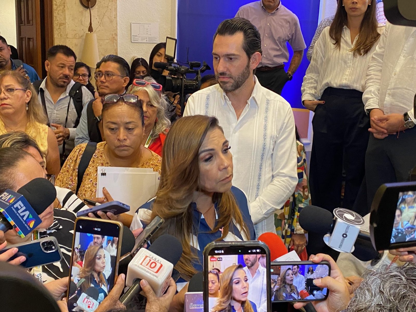 A crowded press conference with a woman speaking while surrounded by reporters and cameras, indicating a tense atmosphere.$# CAPTION
