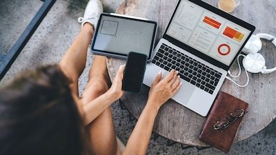 A person using a laptop and phone while working at a table with a coffee and headphones nearby