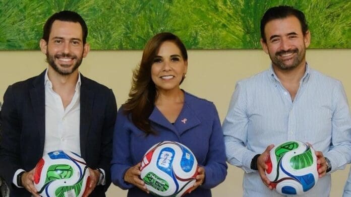 Three individuals stand together holding footballs, smiling, in a well-lit indoor setting with green artwork in the background.$# CAPTION