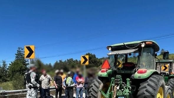 A group of people standing near a tractor at a roadblock under a clear blue sky with road signs indicating a curve ahead.$#$ CAPTION