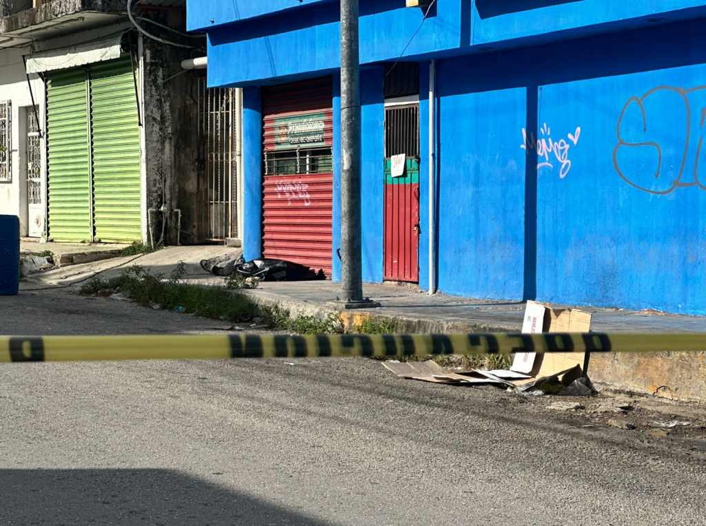 A street scene featuring a red and green storefront with a blue wall, police tape across the foreground, and a pile of trash near the sidewalk.$# CAPTION