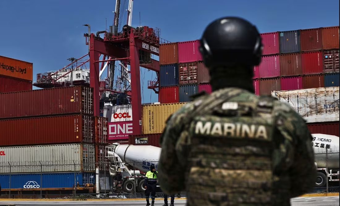 A military personnel in uniform stands watch near shipping containers at a port, with cranes and a truck in the background.