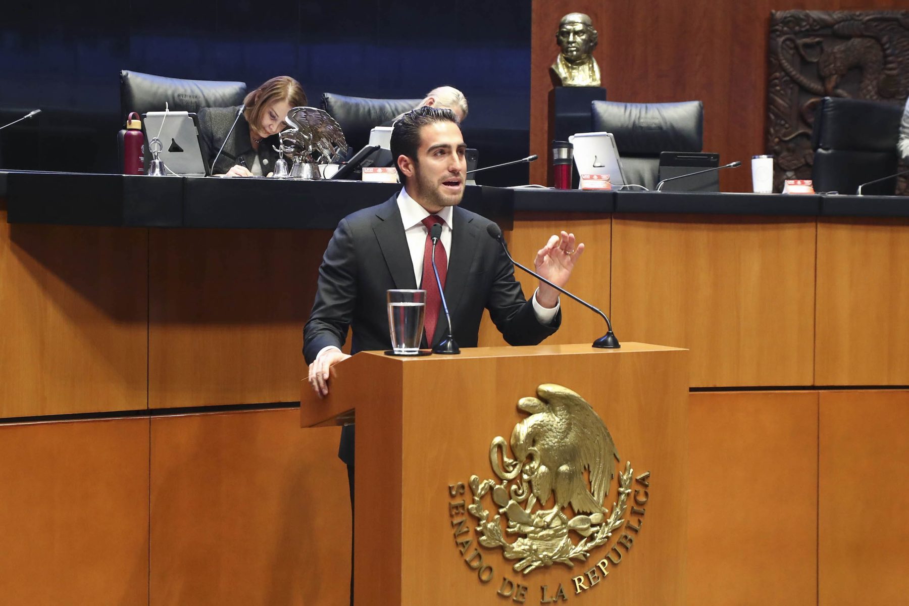 A senator speaking at the podium in a legislative chamber, with colleagues in the background.$# CAPTION