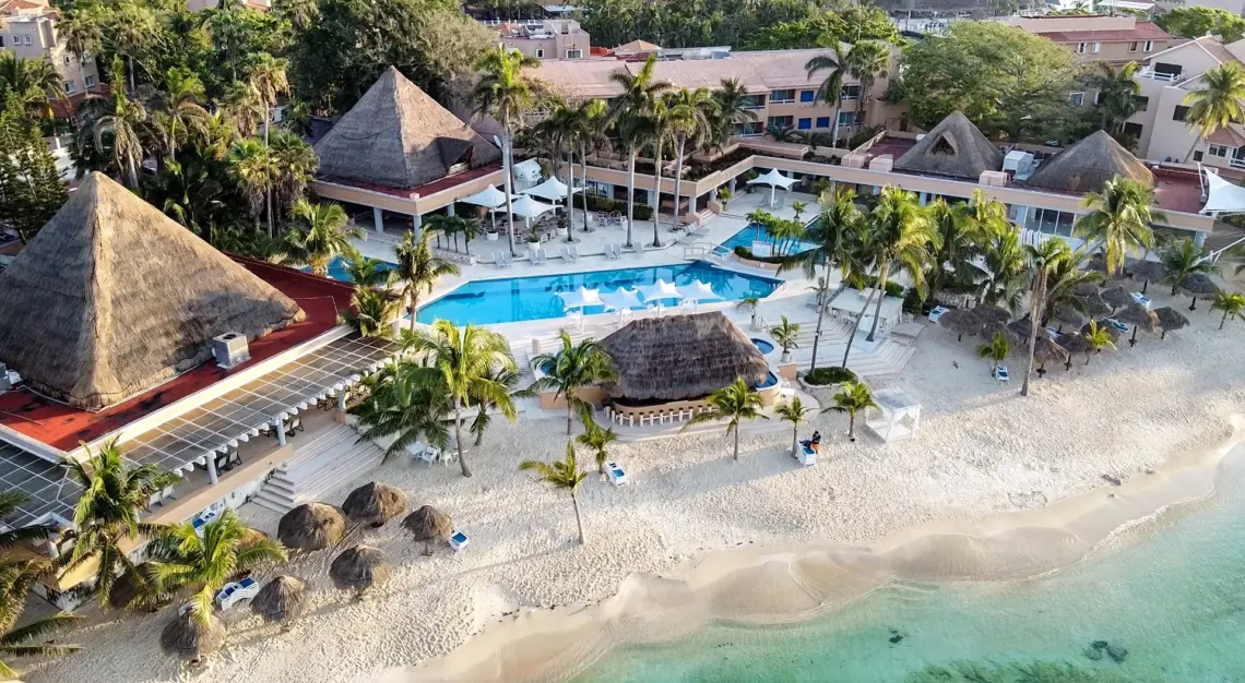 Aerial view of a beachside resort featuring a pool, palm trees, and thatched-roof structures on sandy shores.$#$ CAPTION