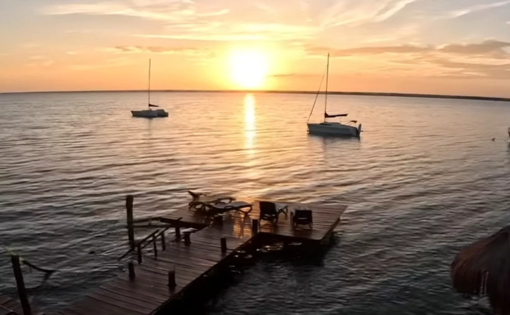 A picturesque sunset over the water, with sailboats in the distance and a wooden dock in the foreground.