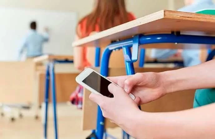 A student sits at a desk holding a smartphone while other students and a teacher are visible in the background of a classroom.$# CAPTION
