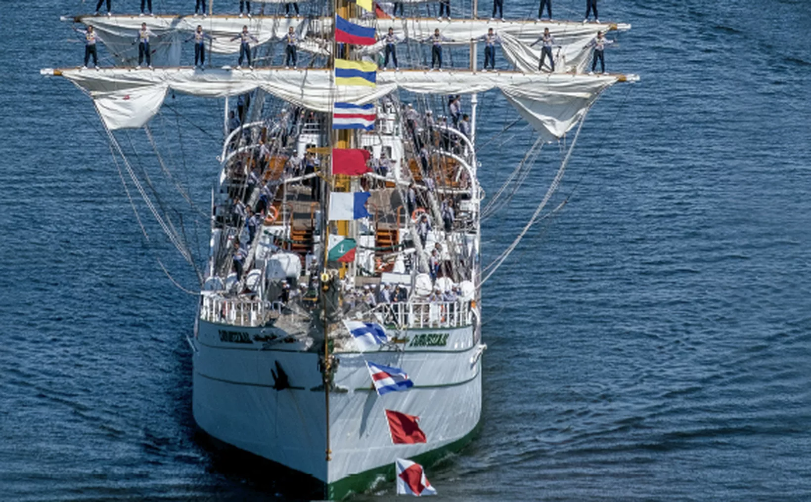 A tall ship with multiple sails and crew members on deck, adorned with colorful flags, sailing on calm waters.$# CAPTION