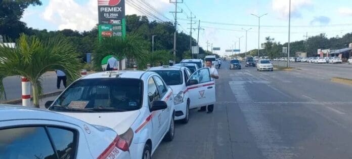 A row of white taxis with red markings parked on the side of a street near a PEMEX gas station, with palm trees and traffic visible in the background.$
