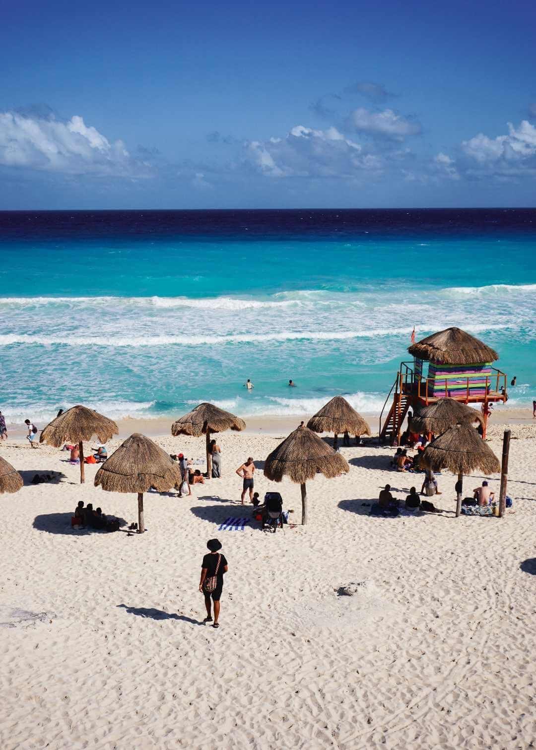 A beautiful beach in Cancun with straw umbrellas, people relaxing, and waves crashing on the shore under a clear blue sky.$# CAPTION