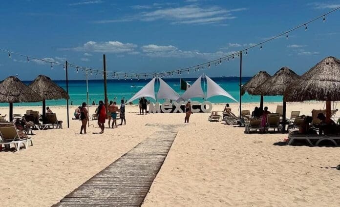 A sandy beach in Mexico with straw umbrellas, lounge chairs, and a clear blue ocean in the background. There are people walking and relaxing along the beach. A large, white decorative tent is set up near the water.$#$ CAPTION
