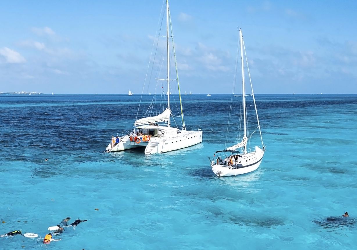 Two sailboats anchored in clear turquoise waters with people enjoying the sea, including snorkelers, in a sunny tropical setting.$# CAPTION