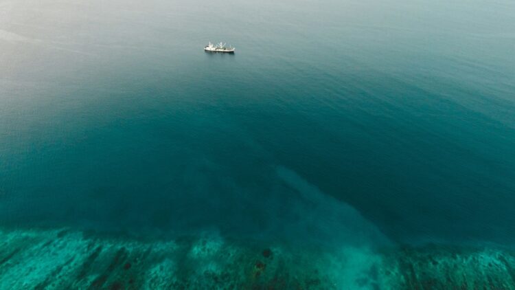 An aerial view of a calm sea with a solitary ship in the distance, showing a gradient of deep blue to turquoise water.