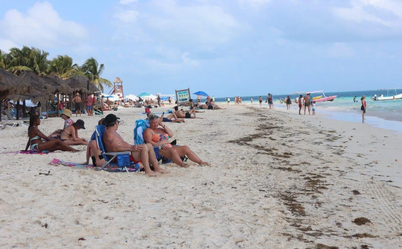 People relaxing on a sandy beach with umbrellas and boats in the background.