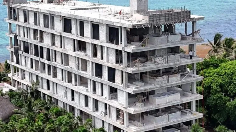 A seven-story concrete building under construction near the coast, with exposed floors and balconies surrounded by greenery and ocean.$#$ CAPTION