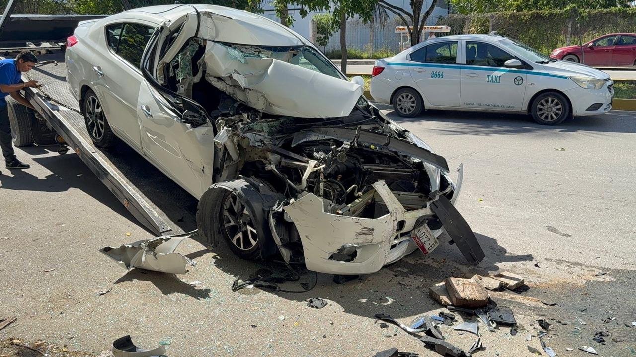 A severely damaged white car on a tow truck, with debris scattered around the scene, and another car parked nearby. A person seems to be inspecting the wreckage.