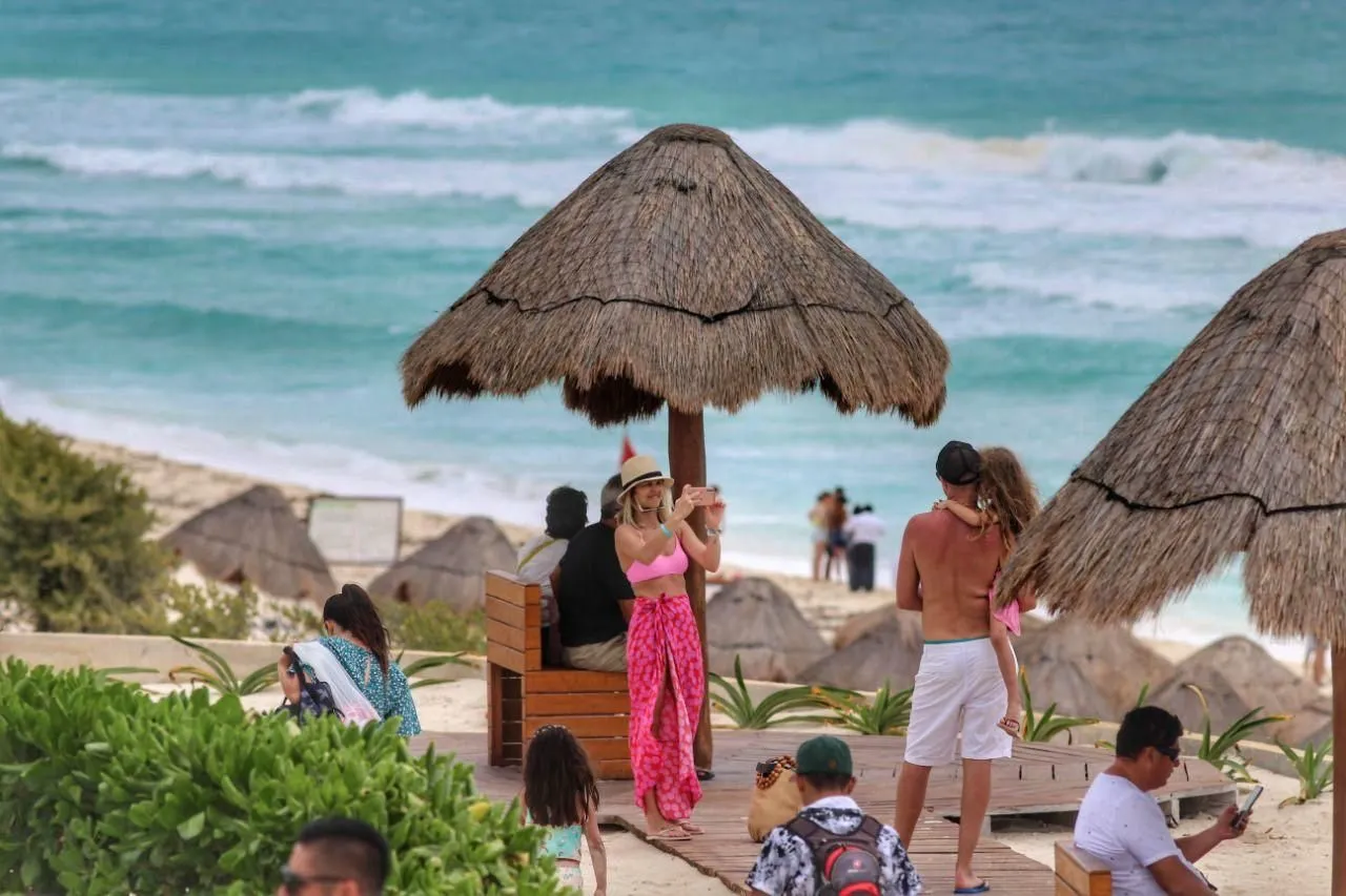 A lively beach scene in Cancun featuring people enjoying the sun, palm trees, and thatched umbrellas near the ocean. A woman in pink takes a photo while others relax and socialize.$#$ CAPTION