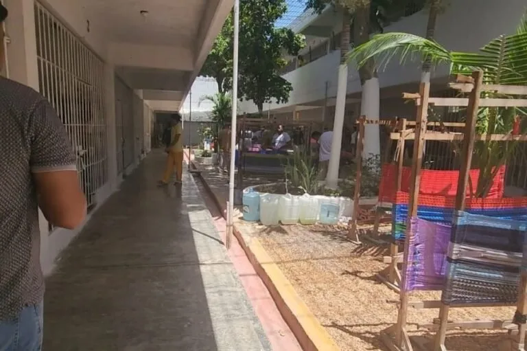 A narrow walkway leads to a colorful courtyard filled with vendors and greenery. Vendors display goods beside large water containers and handwoven textiles. People can be seen engaging in activities in the background.$#$ CAPTION