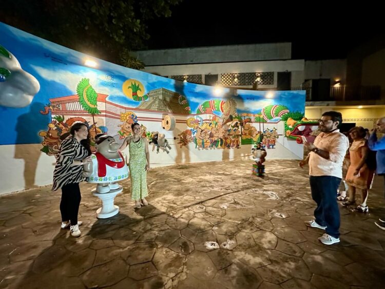 Two individuals posing beside a colorful mural while another person interacts with them in a lively outdoor setting at night.$#$ CAPTION