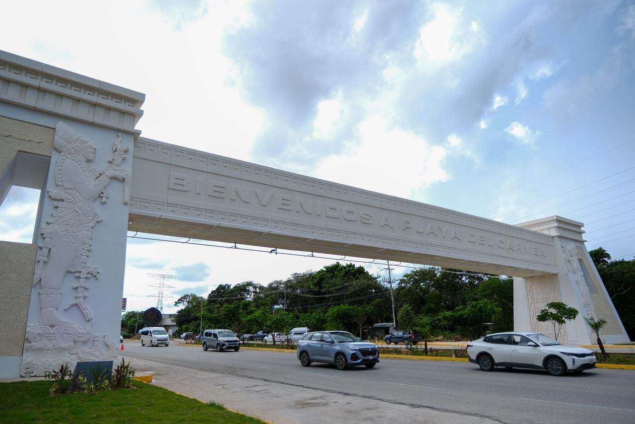 A large white archway with "Bienvenidos a Playa del Carmen" inscribed on it, surrounded by greenery and traffic.$#$ CAPTION