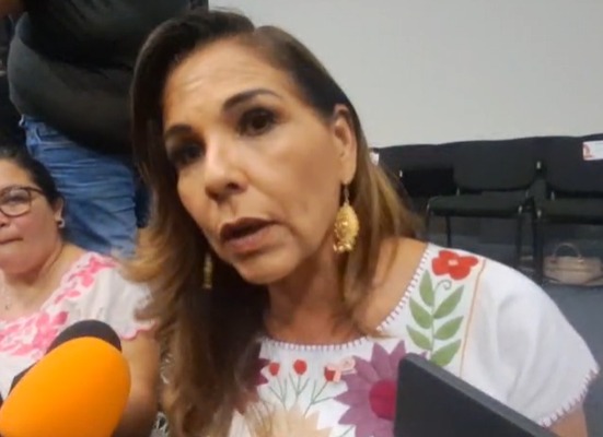 a woman with long hair and earrings speaking in front of a microphone with a person in the background-$# CAPTION