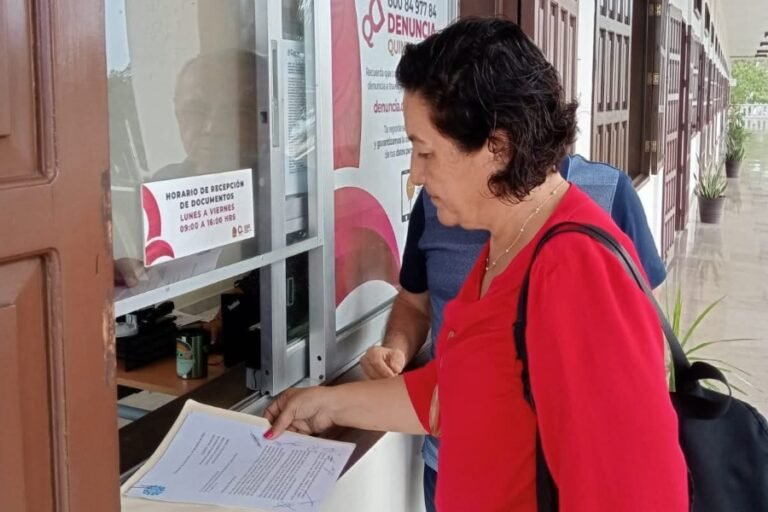 A woman in a red shirt is handing over documents at a service window while another person stands behind her. The window displays information about document submission hours in Spanish.