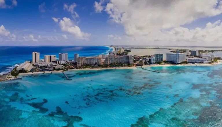 A scenic aerial view of a vibrant turquoise sea, sandy beach, and coastal buildings under a blue sky with clouds.$# CAPTION