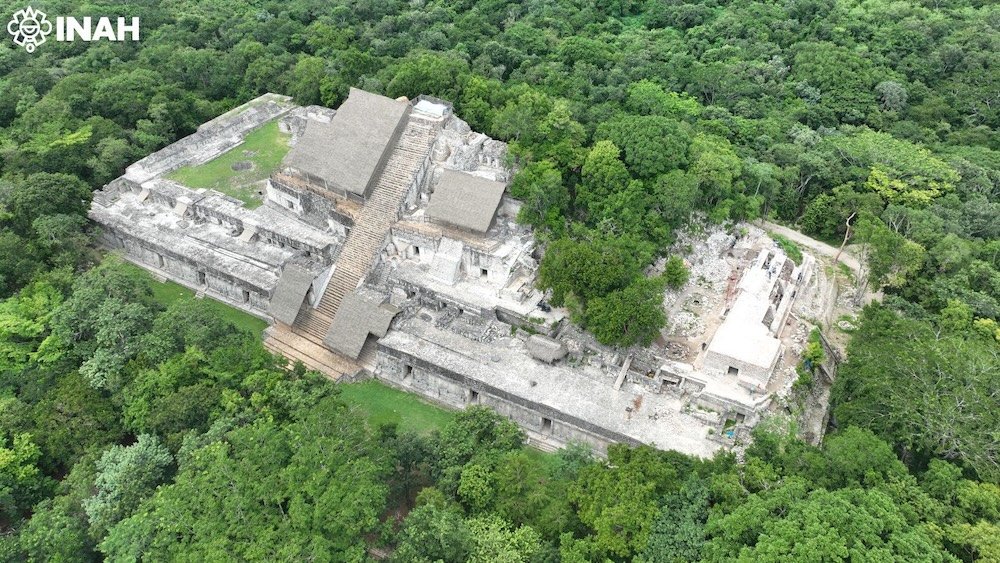 Aerial photograph of ancient Mayan ruins surrounded by dense greenery, showcasing stone structures and stairways.$#$ CAPTION
