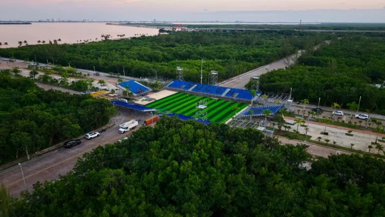 An aerial view of a sports field with blue bleachers, surrounded by lush greenery and a body of water in the background.$#$ CAPTION